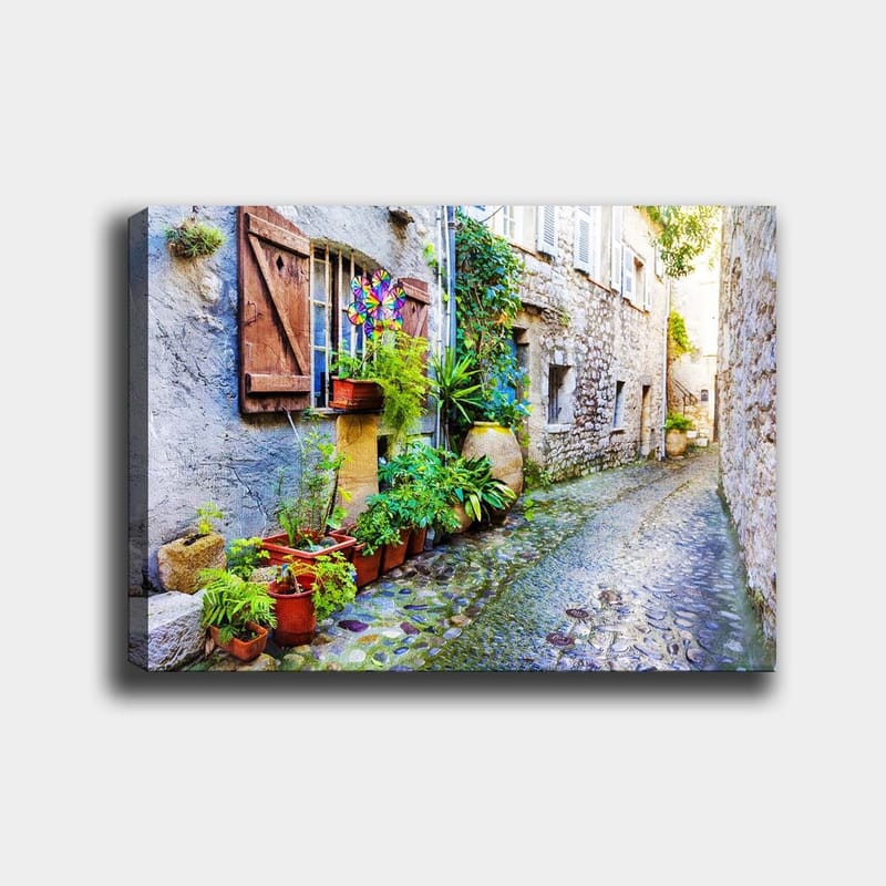 Lærred 70x100 cm - Charmerende gyde med stenhuse og blomsterkrukker skaber en livlig atmosfære, Grøn / Blå / Beige