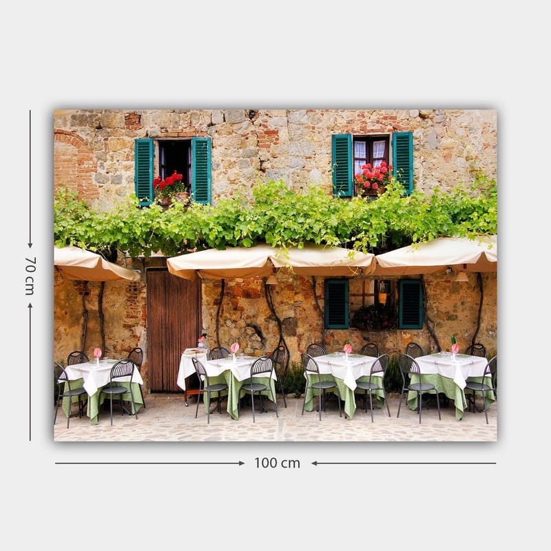 Lærred 70x100 cm - Charmerende udendørs spiseplads med hvide borde og grønne duge foran en gammel stenbygning med mørkegrønne skodder - Mørkegrøn / Hvid / Teglrød - Boligtilbehør - Billeder & kunst - Billeder på lærred