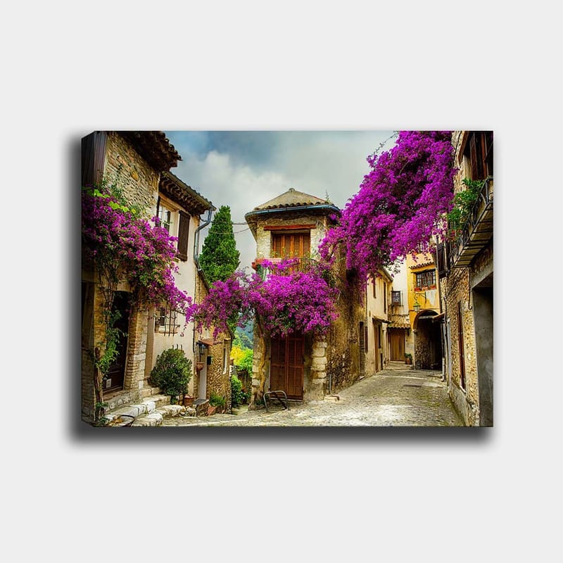 Lærred 70x100 cm - Malerisk gyde omgivet af farverige blomster i charmerende landsbyomgivelser, Lilla / Grøn / Beige