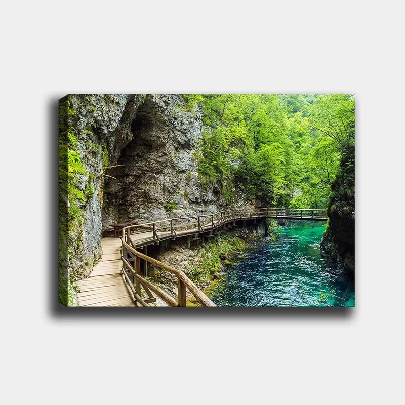 Lærred 70x100 cm - Naturskøn udsigt over en træbro, der snor sig gennem en frodig kløft ved en klar, turkis flod, Grøn / Turkis / Grå