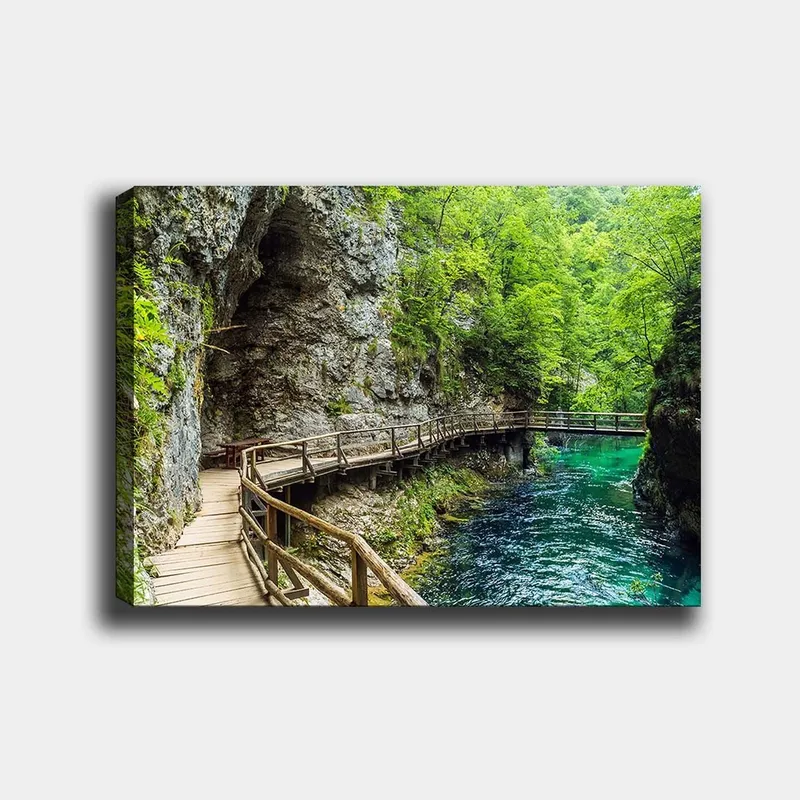 Lærred 70x100 cm - Naturskøn udsigt over en træbro, der snor sig gennem en frodig kløft ved en klar, turkis flod, Grøn / Turkis / Grå