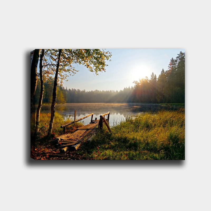 Lærred 70x100 cm - Rolig sø med en træbro omgivet af frodige træer og morgensolens stråler, Grøn / Brun / Gul