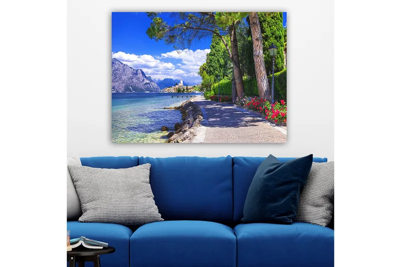 Lærred 70x100 cm - Smuk promenade med træer og blomster ved en sø, omgivet af bjerge og en malerisk by i baggrunden - Blå / Grøn / Rød - Boligtilbehør - Billeder & kunst - Billeder på lærred