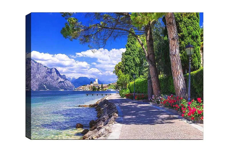 Lærred 70x100 cm - Smuk promenade med træer og blomster ved en sø, omgivet af bjerge og en malerisk by i baggrunden, Blå / Grøn / Rød