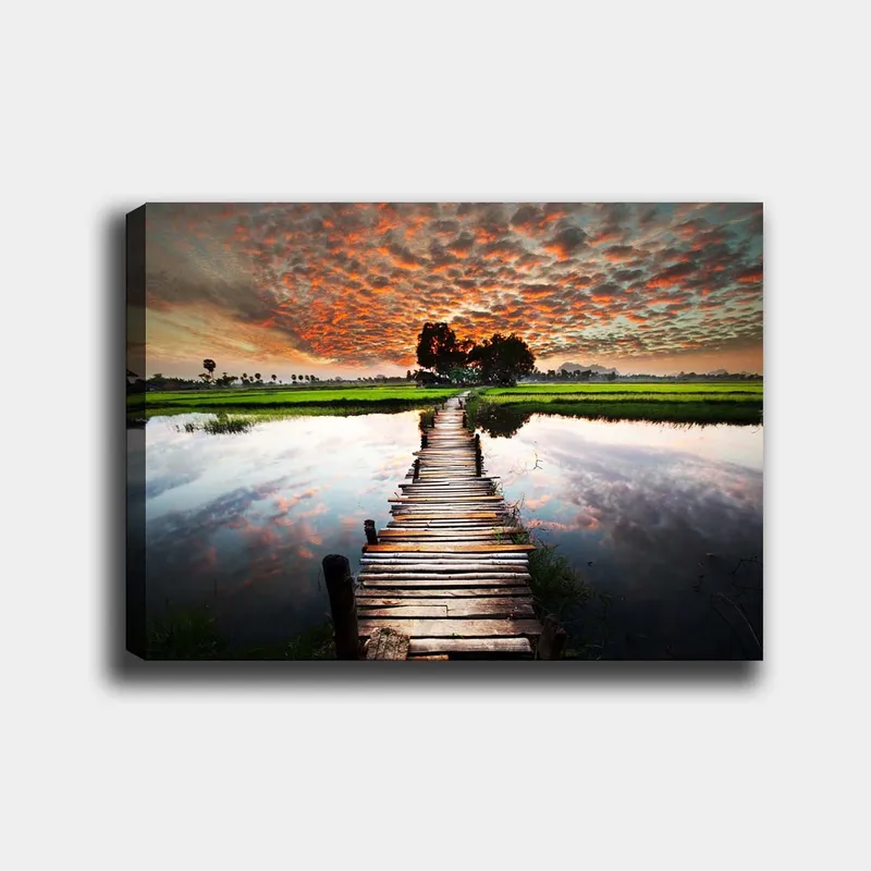 Lærred 70x100 cm - Træbro, der spænder over en spejlblank vandoverflade, omgivet af frodigt grønt og en dramatisk solnedgang, Orange / Grøn / Blå