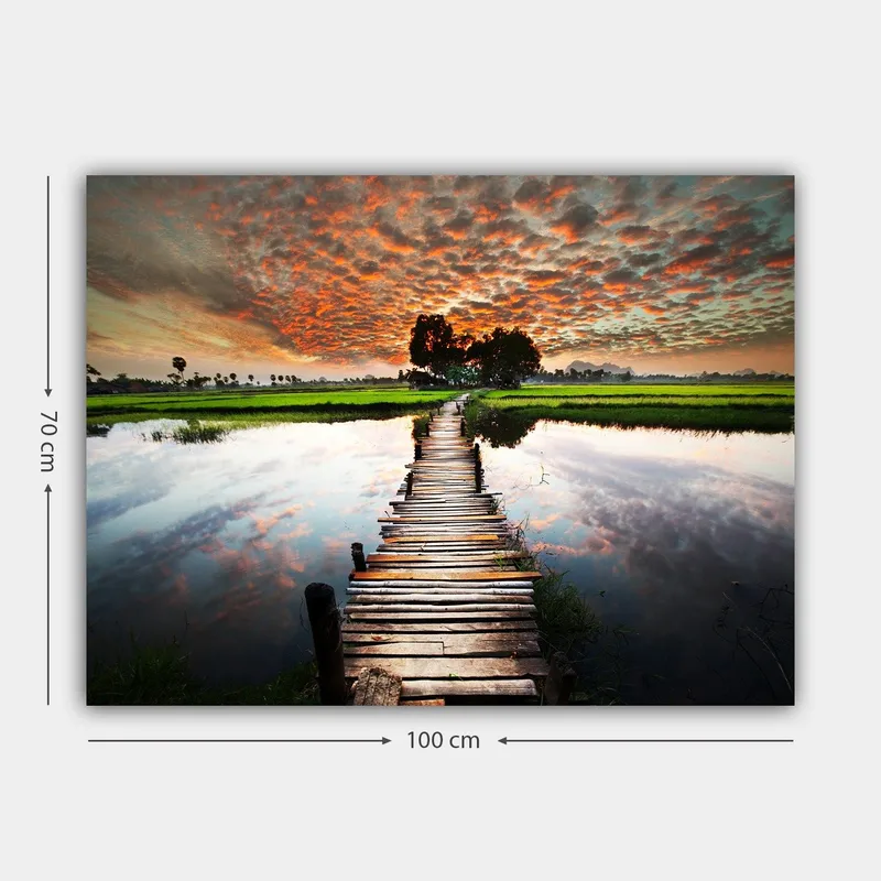 Lærred 70x100 cm - Træbro, der spænder over en spejlblank vandoverflade, omgivet af frodigt grønt og en dramatisk solnedgang - Orange / Grøn / Blå - Boligtilbehør - Billeder & kunst - Billeder på lærred